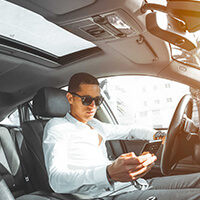 A man wearing a white top and sunglasses in his car checking his phone
