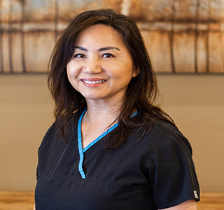 A female hygienist with brunette hair  wearing blue scrubs, background is a painting of trees